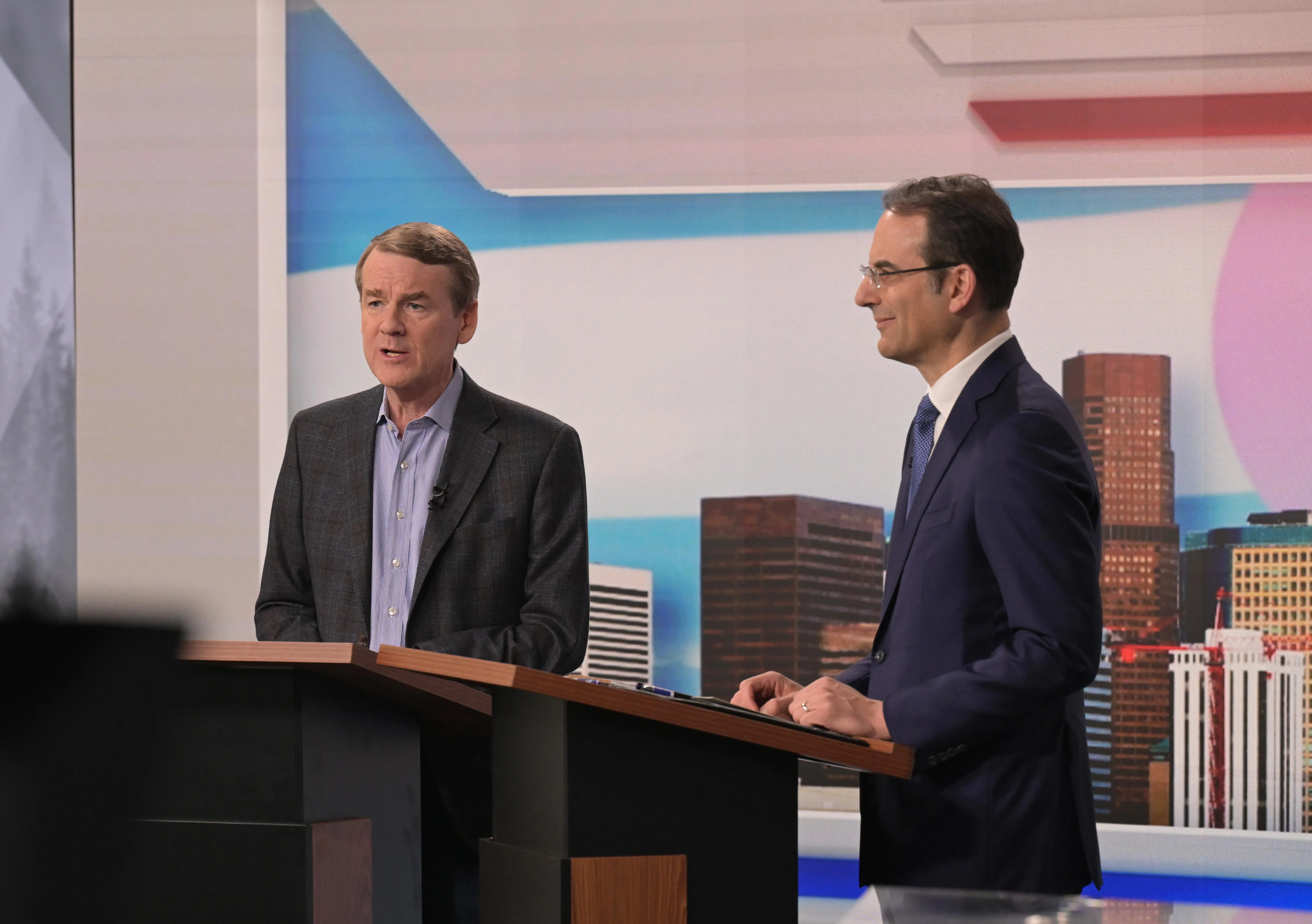 Sen. Michael Bennet, left, and Attorney General Phil Weiser stand behind separate podiums at a TV studio