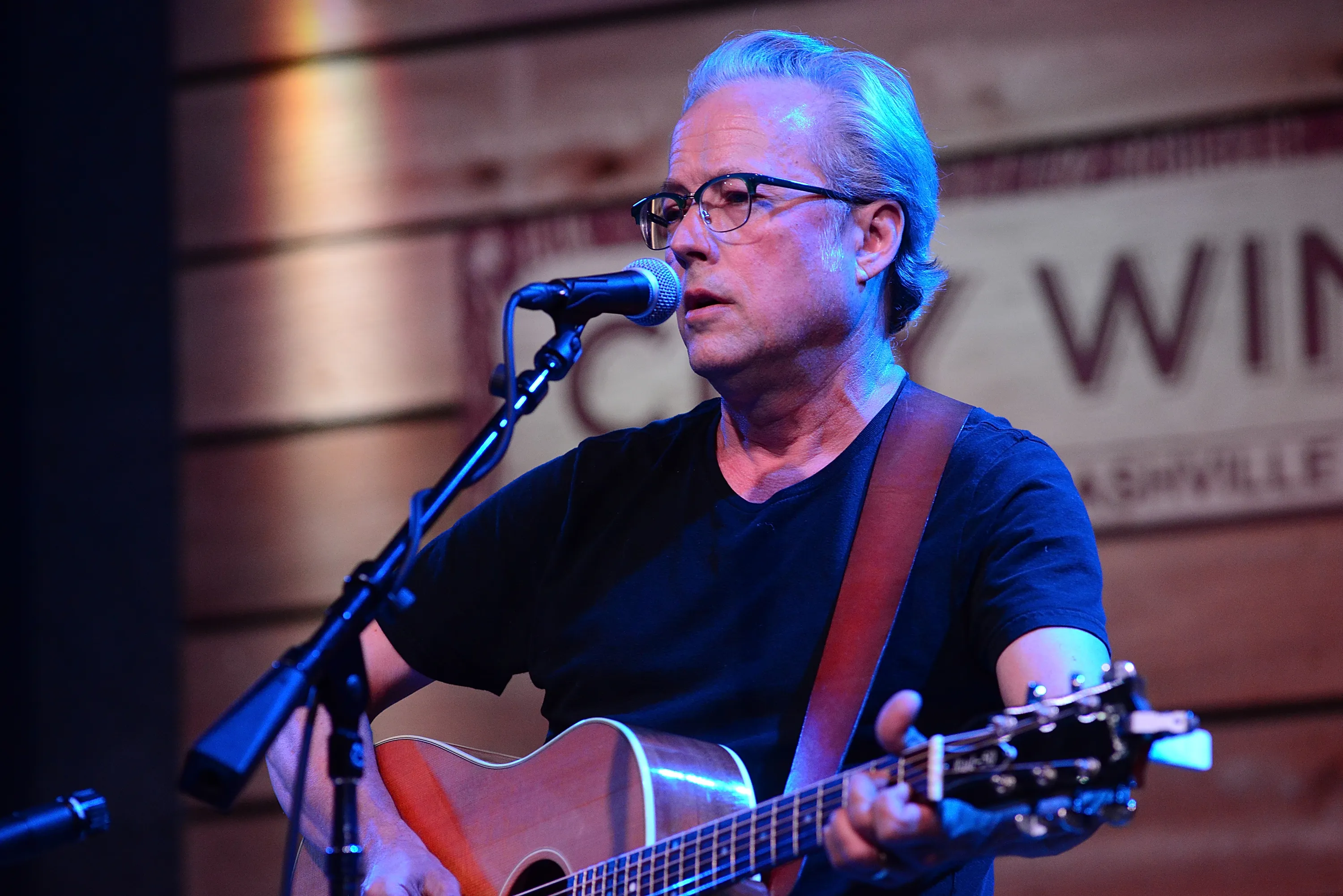 A man with glasses and grey hair sings into a microphone while playing an acoustic guitar.