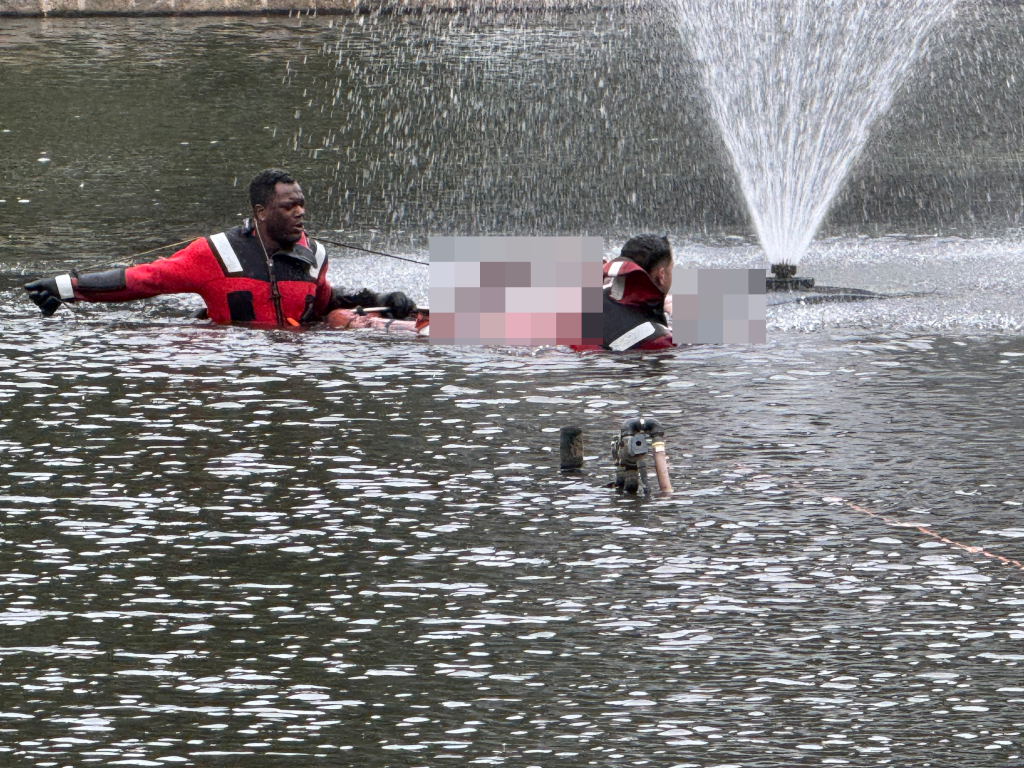 A rescuer in a red and black wetsuit pulling a person on a floating stretcher out of the water near a fountain.