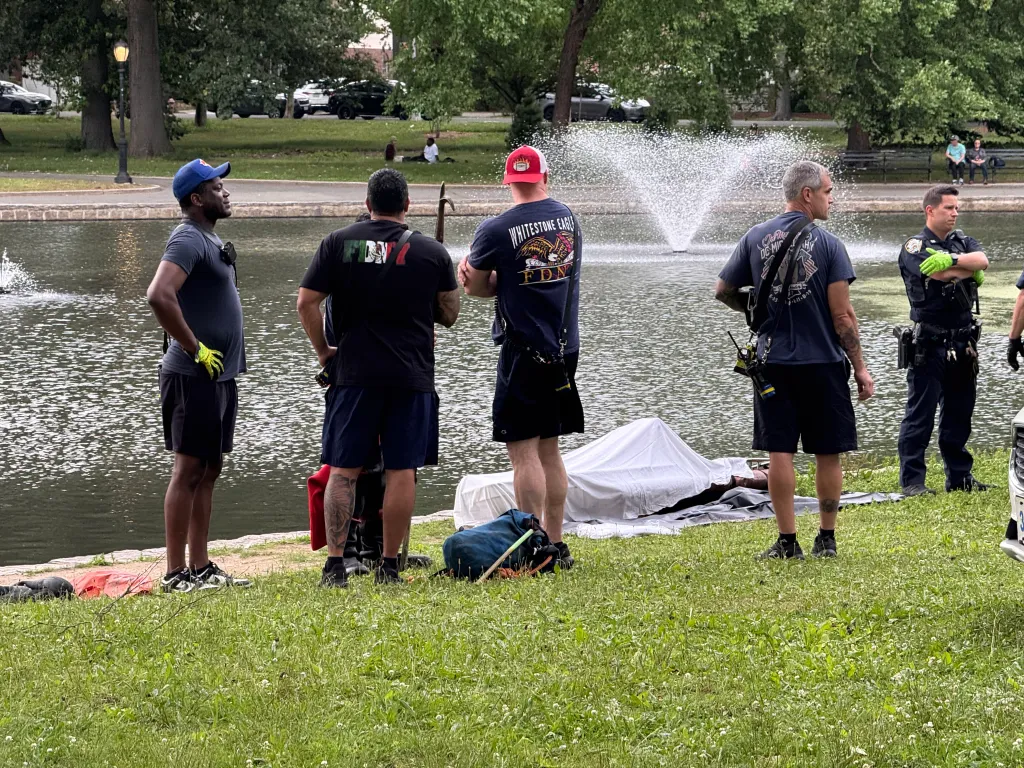 Police and first responders investigate a body covered by a white sheet next to a pond.