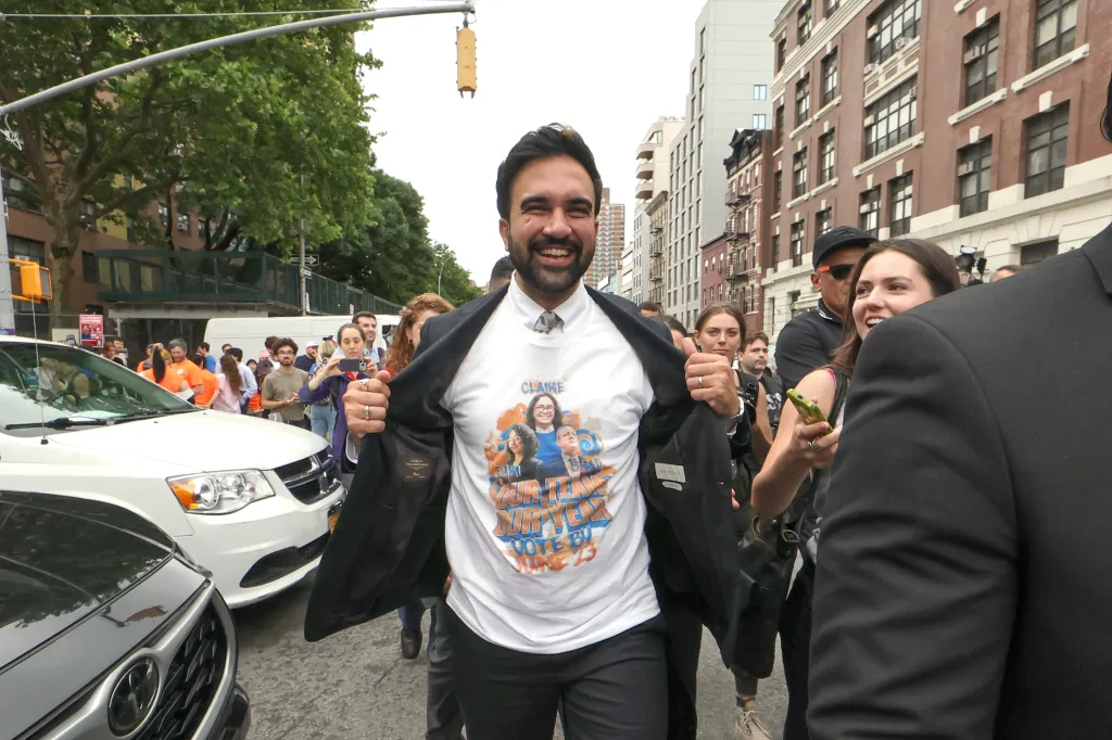 Mayor Zohran Mamdani, smiling, pulls open his suit jacket to reveal a white t-shirt.