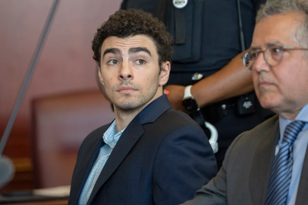 Luigi Mangione, facing a murder charge, looks on while a man in uniform stands behind him, and another man in a suit sits beside him.