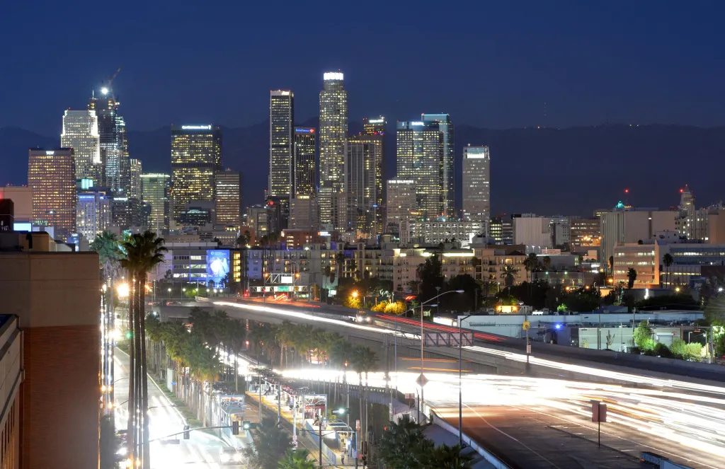 Nighttime view of Los Angeles skyline with downtown buildings lit up and blurred streaks of light from traffic on a highway.