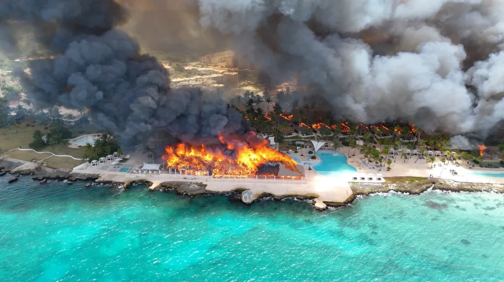 Aerial view of a resort in the Dominican Republic with a building engulfed in flames and palm trees burning, emitting large plumes of dark smoke.