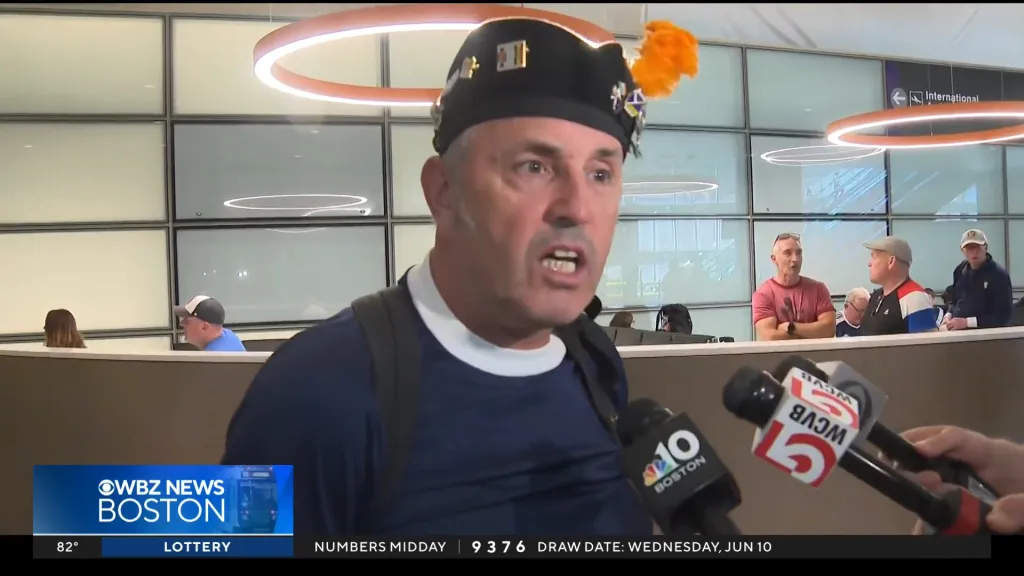 A man in a kilt-style hat with an orange plume speaks into news microphones.