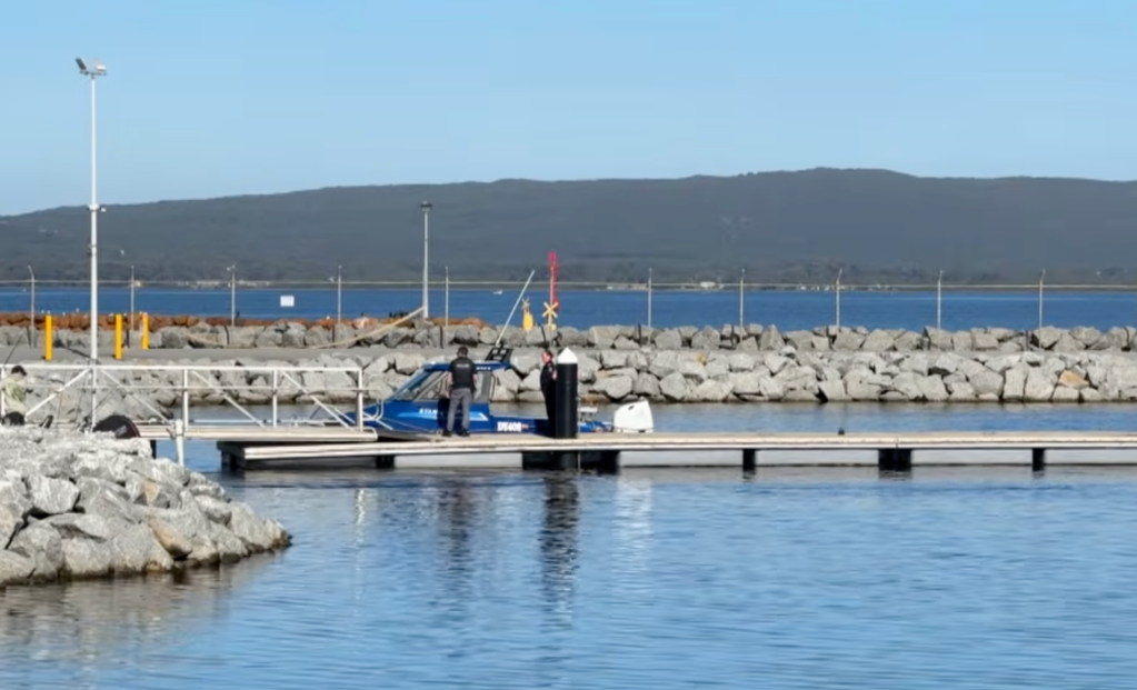 A blue boat docked at a pier, with a person standing on it and another on the pier.
