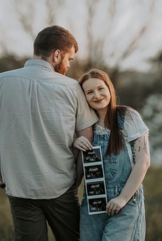 A man standing with his back to the viewer, and a woman leaning on his shoulder holding ultrasound pictures, smiling at the camera.n