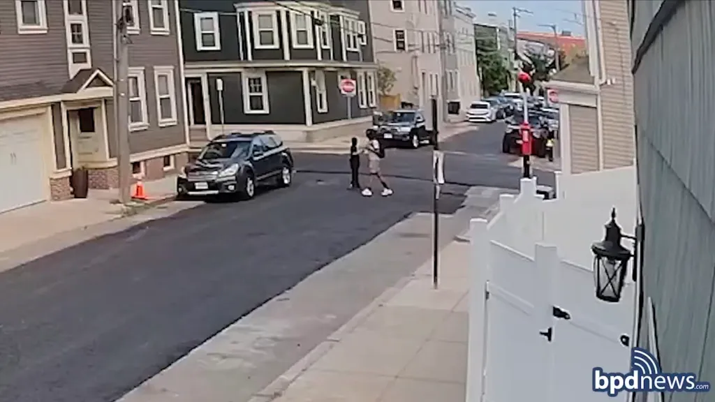 A surveillance camera captures two individuals interacting with a vehicle on a residential street.