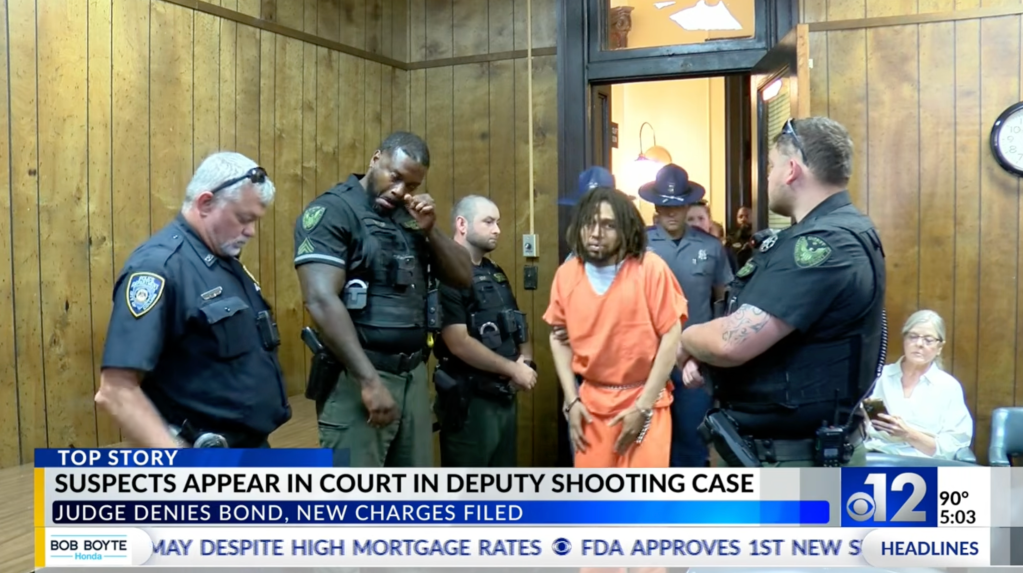 A man in an orange jumpsuit is escorted by officers in a courtroom, with text 