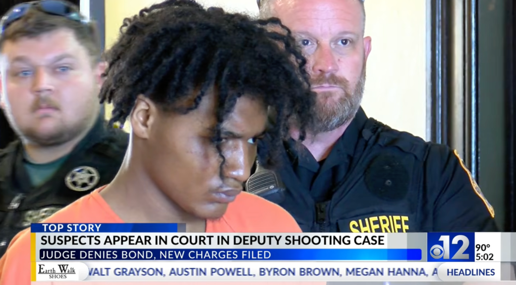 A man in an orange jumpsuit with dark dreadlocks appears in court, flanked by two sheriffs, as a news chyron reads, 