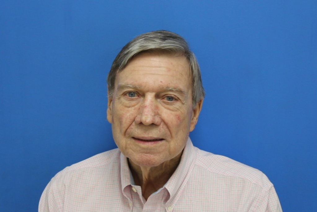 Headshot of an older man with gray hair and a pink plaid shirt against a blue background.