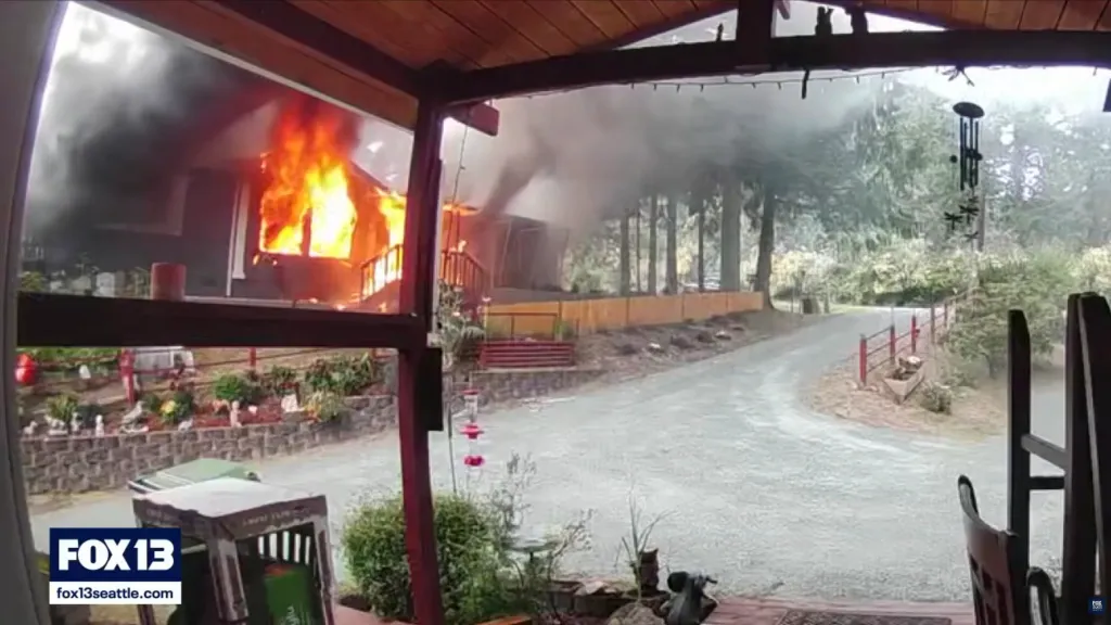 A house engulfed in flames, with thick smoke rising, viewed from a porch.