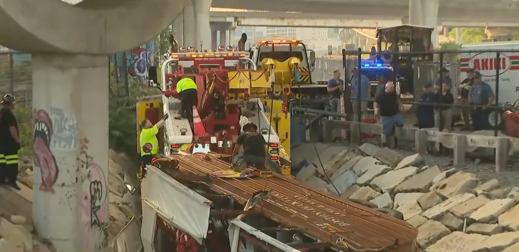 A large yellow tow truck working to pull a brown train car out from under a bridge.