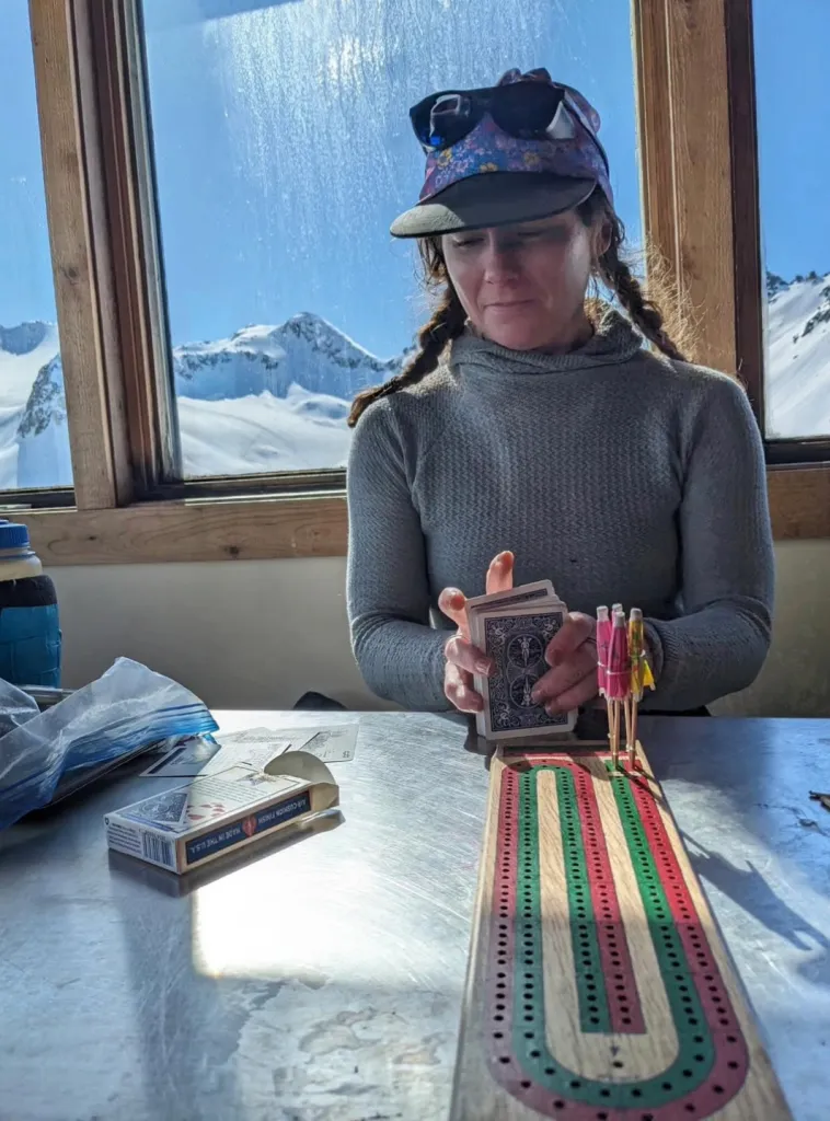 A woman playing cards and cribbage inside a cabin with snowy mountains visible through the window.
