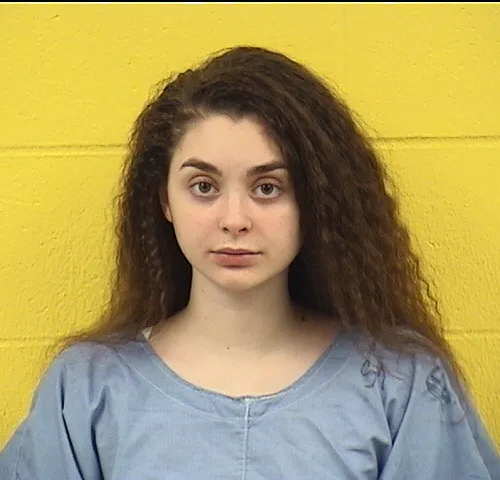 A mugshot of a young woman with long, curly brown hair wearing a light blue smock.