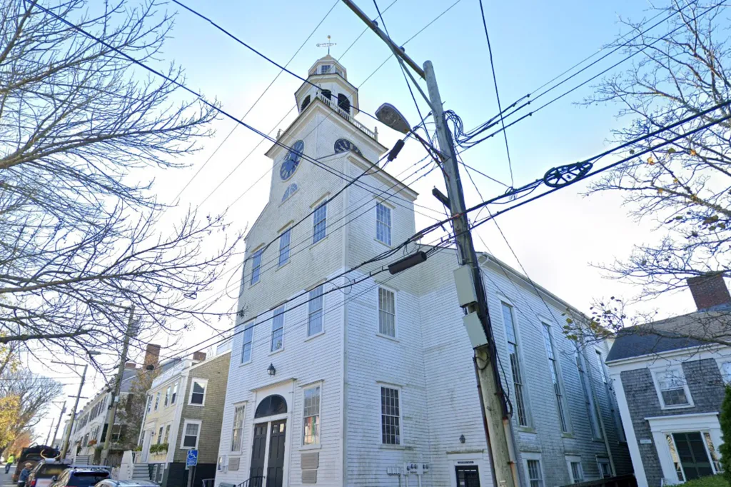 A tall white church with a clock tower and steeple is seen from a low angle, with power lines and bare tree branches crisscrossing the sky.