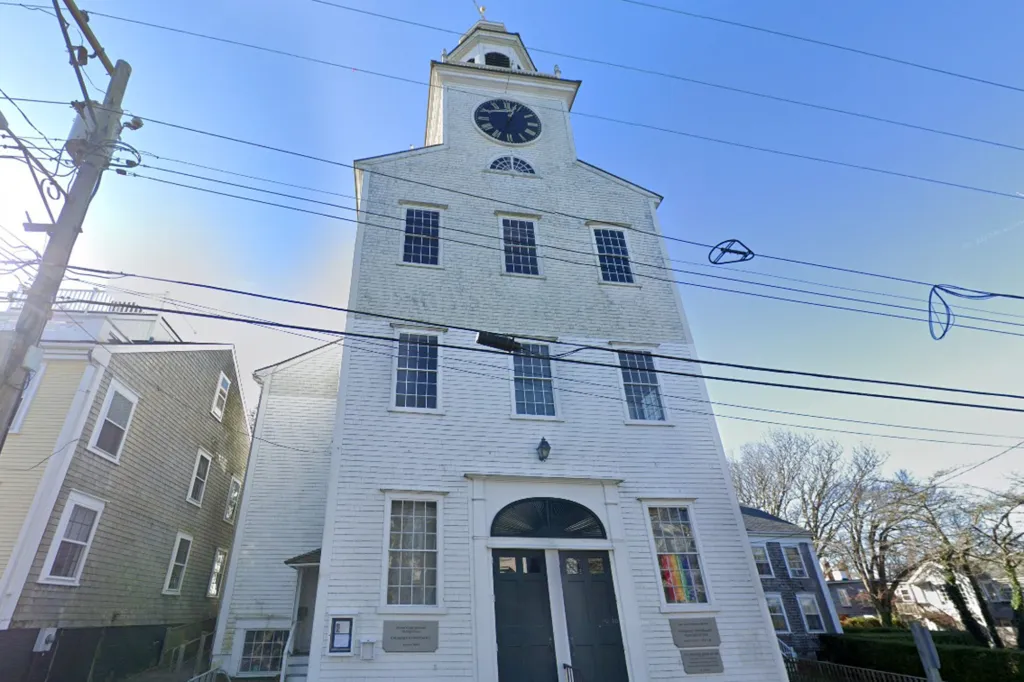 A tall white wooden church building with a clock tower and a rainbow flag in a window.