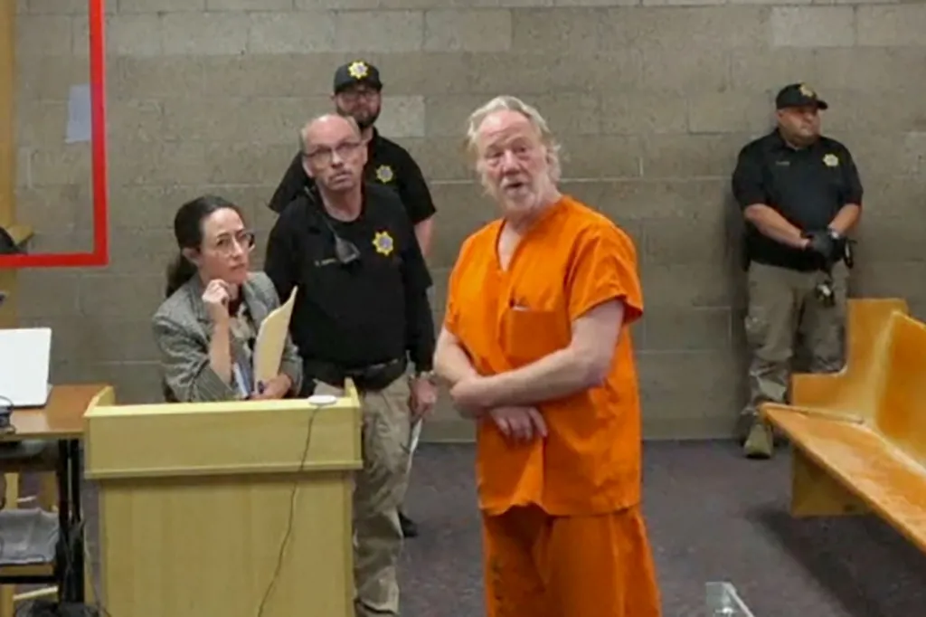 Timothy Busfield in an orange jumpsuit appears in court, flanked by officers, with a woman at a podium.