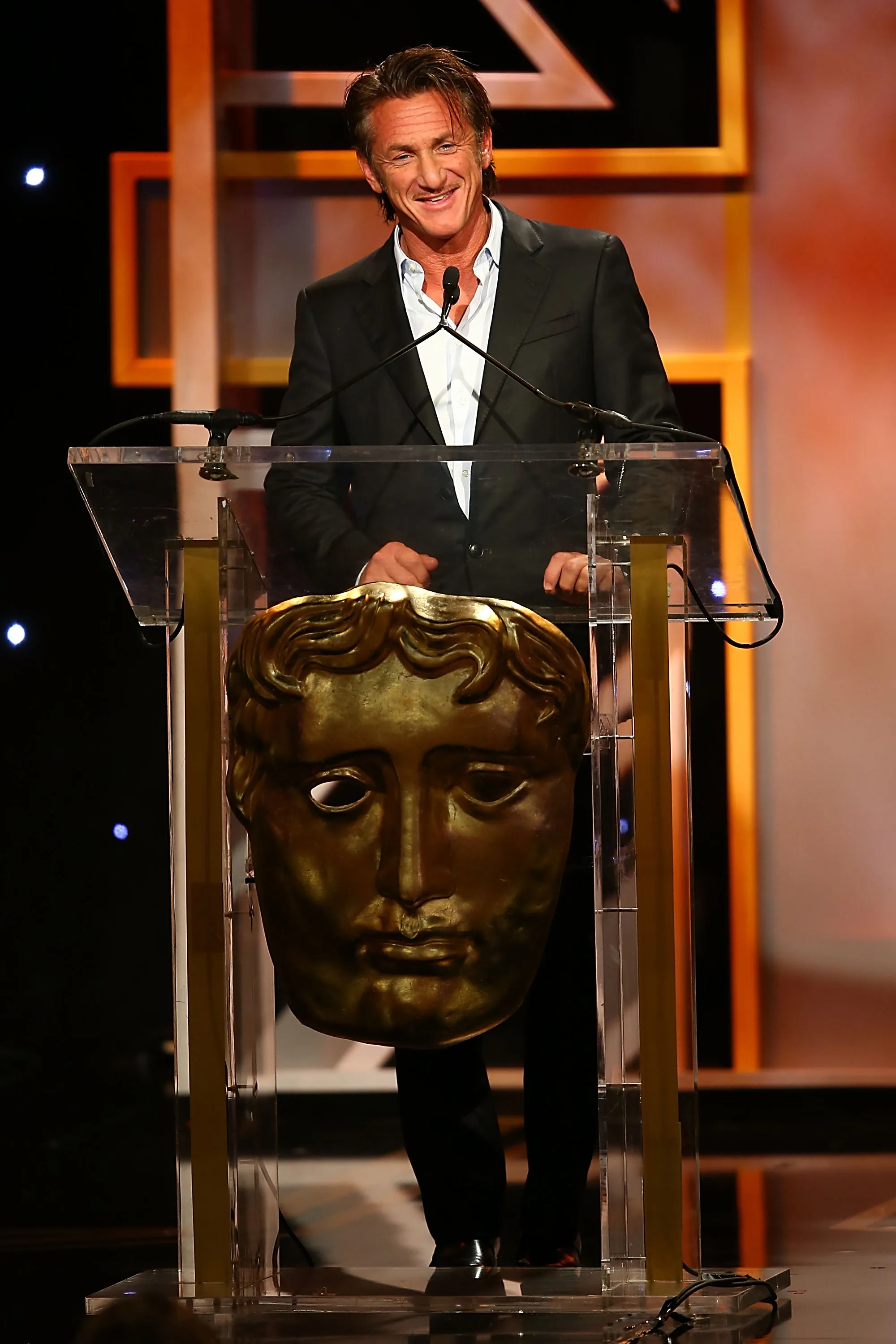 Sean Penn speaking at an awards ceremony behind a podium with a large, golden, mask-like award in front.