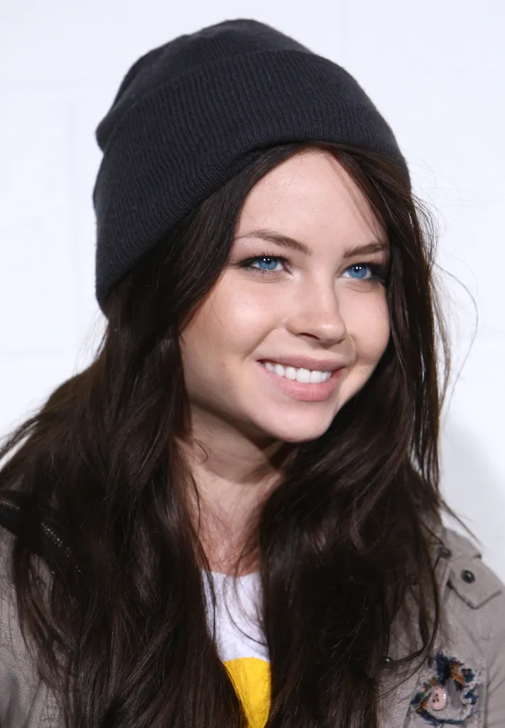 Actress Daveigh Chase smiles, wearing a dark beanie and a light-colored jacket over a white and yellow shirt.
