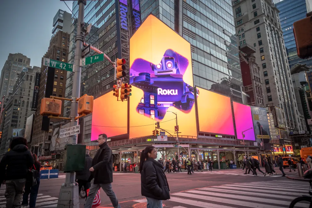 Advertising for Roku in Times Square, New York, with a prominent digital billboard featuring the Roku logo and a robot mascot against a vibrant orange and purple background, surrounded by other digital signs and buildings, and with pedestrians crossing a street in the foreground.