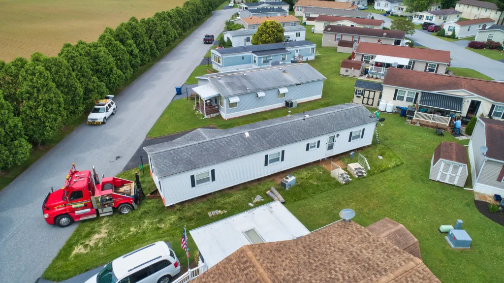 Aerial view of a red tow truck preparing to remove a white manufactured home from a park with other mobile homes.