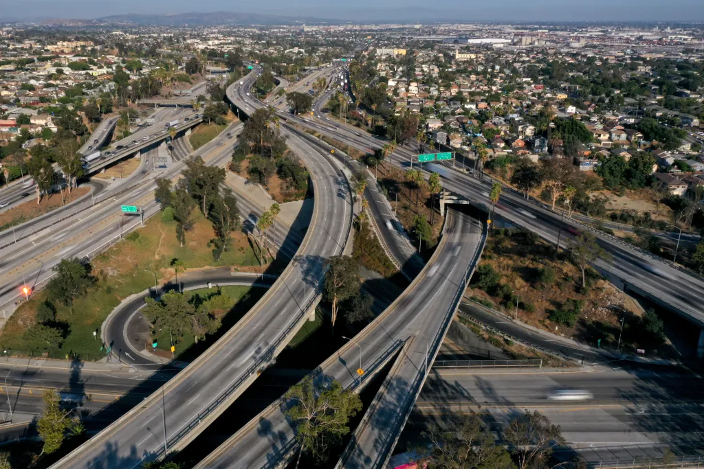 An aerial view of the East Los Angeles Interchange complex, showing multiple highways converging and cars moving, surrounded by a city landscape.
