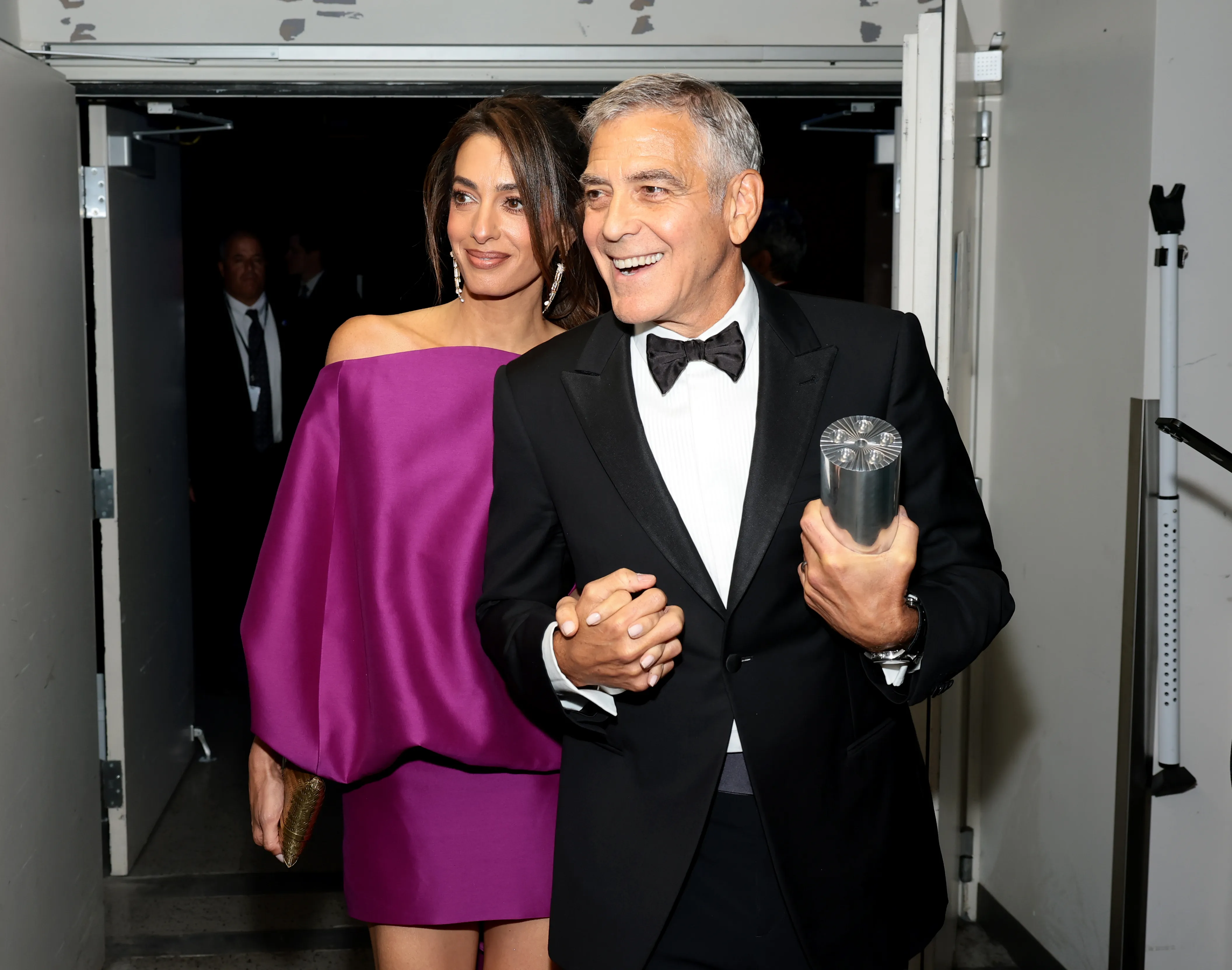 Amal Clooney in a magenta dress and George Clooney in a tuxedo holding an award.