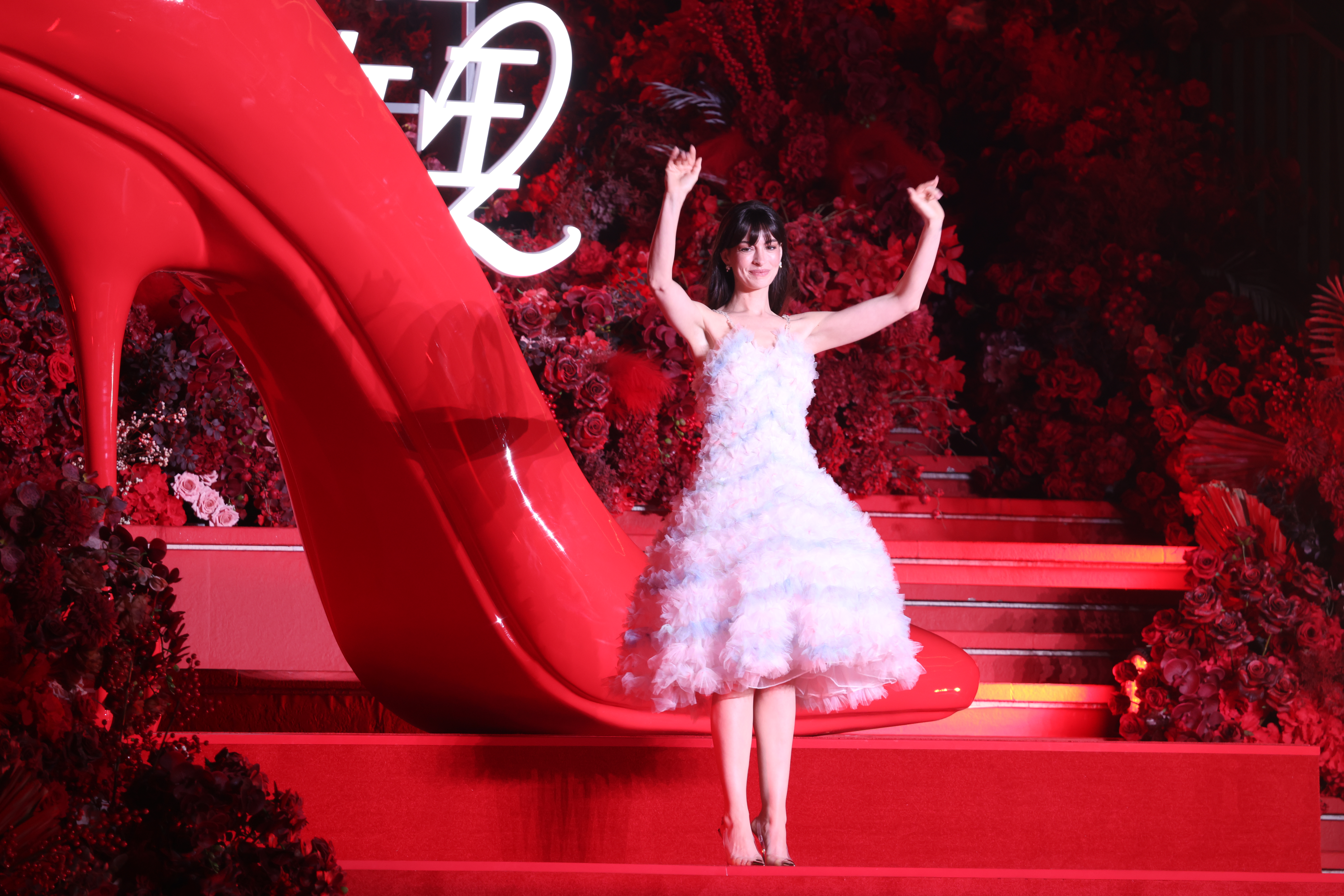 Anne Hathaway in a ruffled white gown, standing next to a large red high heel shoe sculpture.