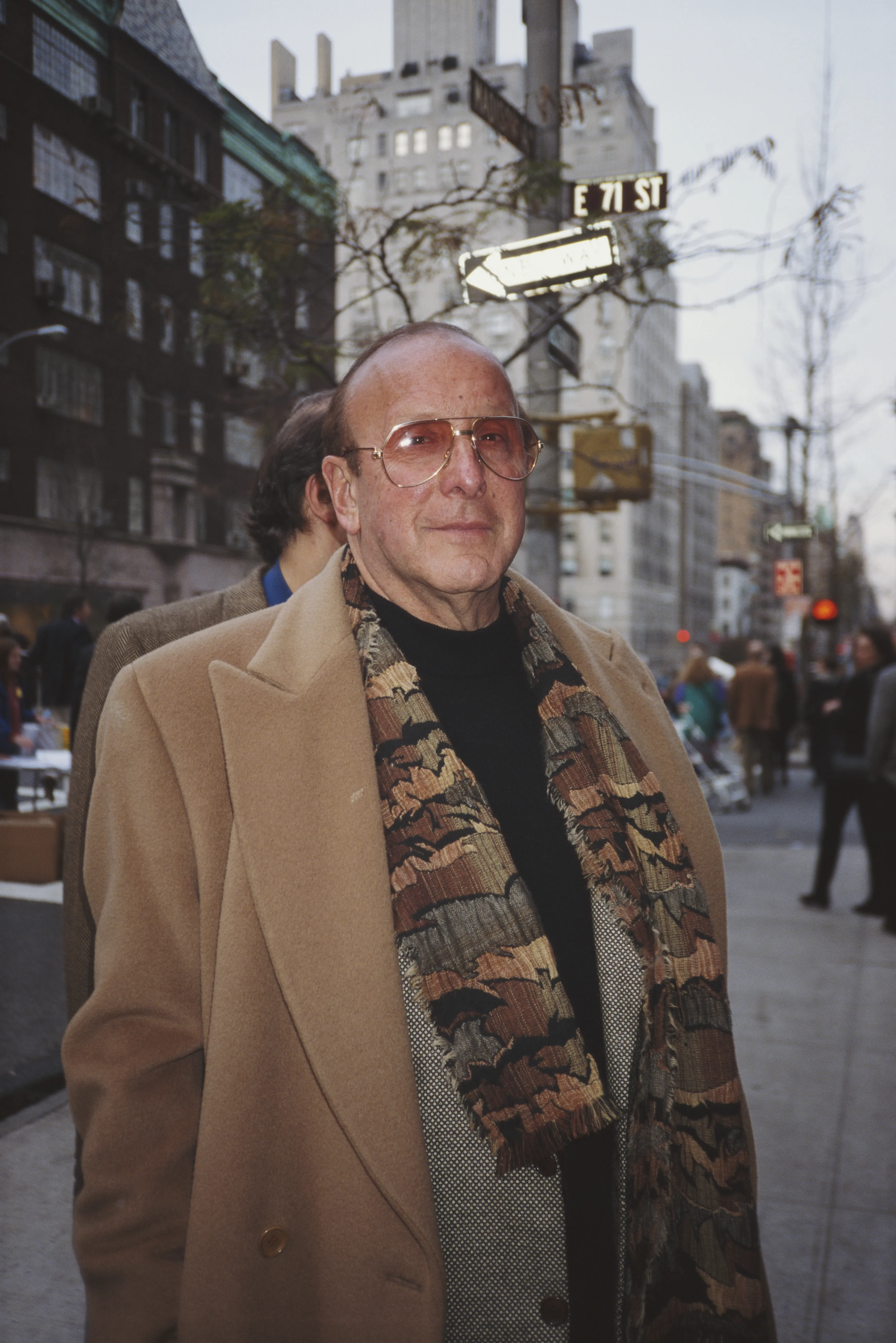 American record producer Clive Davis on a street corner, wearing a camel-colored coat, a black turtleneck, and a patterned scarf.