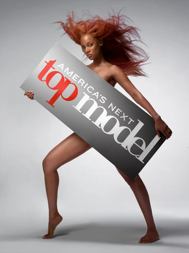 Tyra Banks, with red hair, poses nude while holding a sign that says 