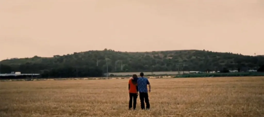 A couple stands in a field with their backs to the viewer, looking at a distant hill.