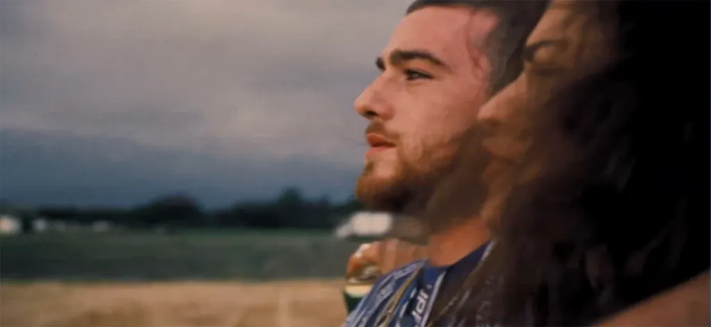 Angus Cloud with a beard, looking to the side with an indistinct figure next to him, with a field and cloudy sky in the background.