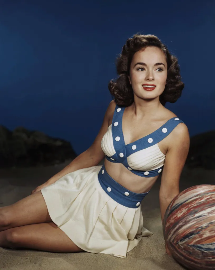 Portrait of Ann Blyth sitting on sand in a two-piece swimsuit.