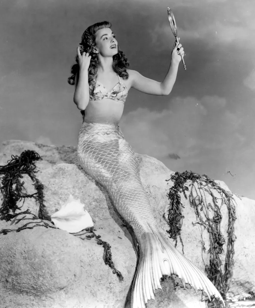 Ann Blyth as a mermaid, adjusting her hair with one hand and holding a mirror with the other, on a rock in 