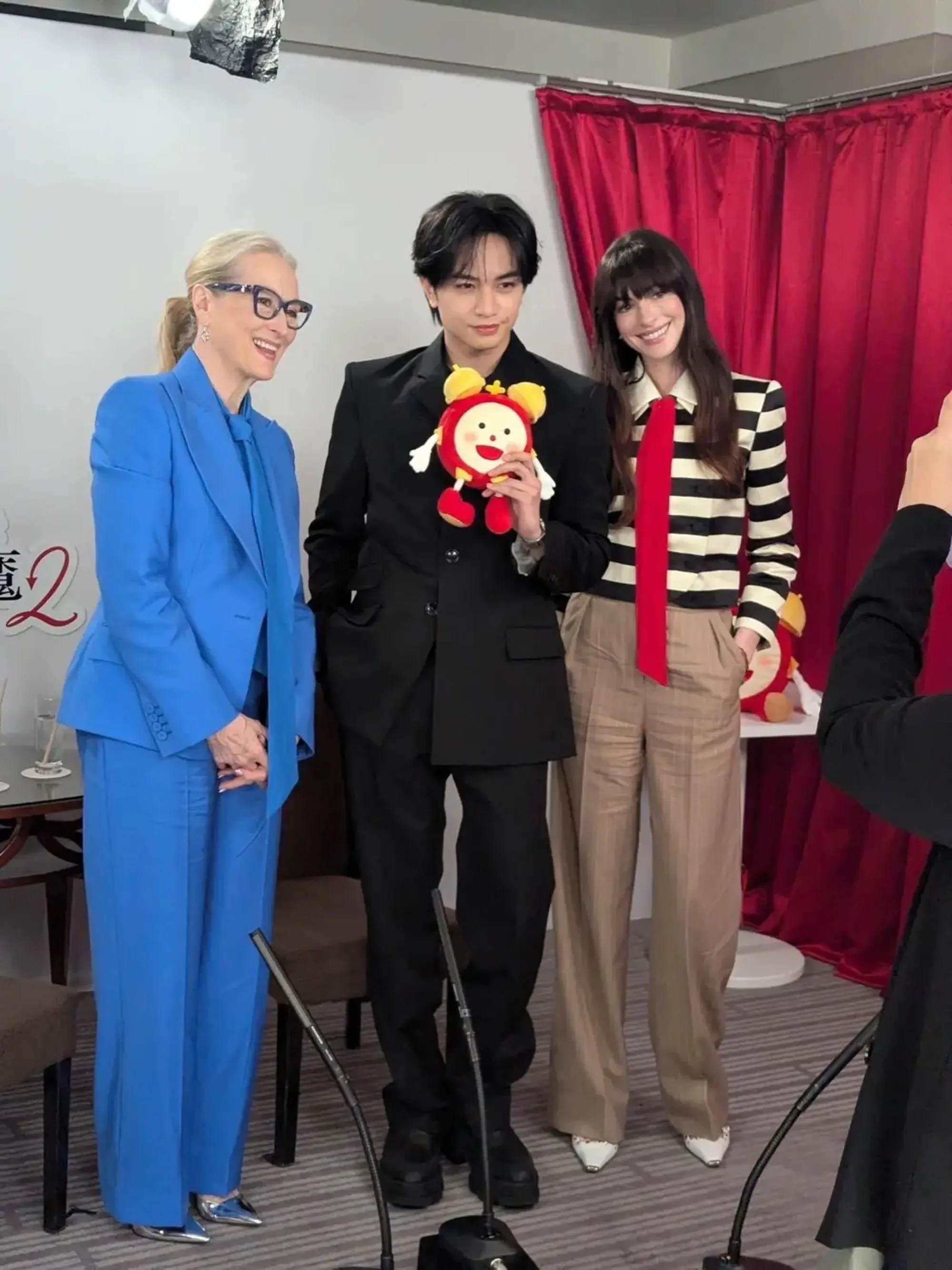 Meryl Streep in a blue suit, a young man in a black suit holding a red plush toy, and Anne Hathaway in a striped top and red tie, standing together in front of red curtains.