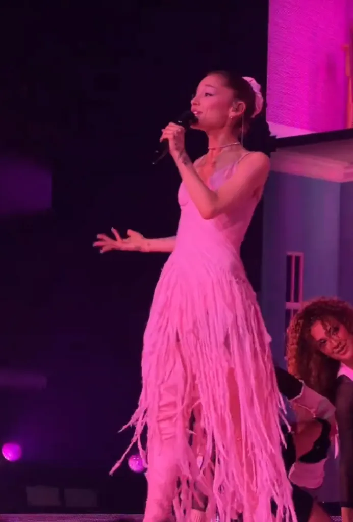 Ariana Grande singing on stage in a long pink fringed dress, with a dancer visible to her right.