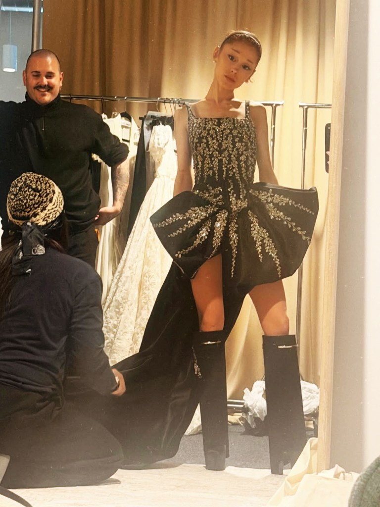 Ariana Grande trying on a black and silver gown in a dressing room, with two people helping her.
