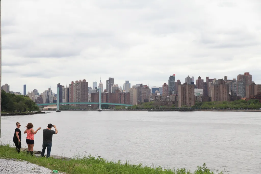 Three people standing by the water, looking at the city skyline with a bridge in the distance.