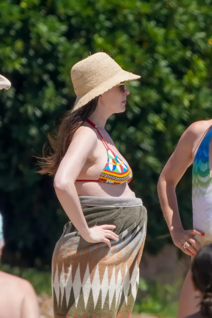Anne Hathaway in a colorful bikini top, straw hat, and sunglasses, with a towel wrapped around her midsection, revealing a baby bump.