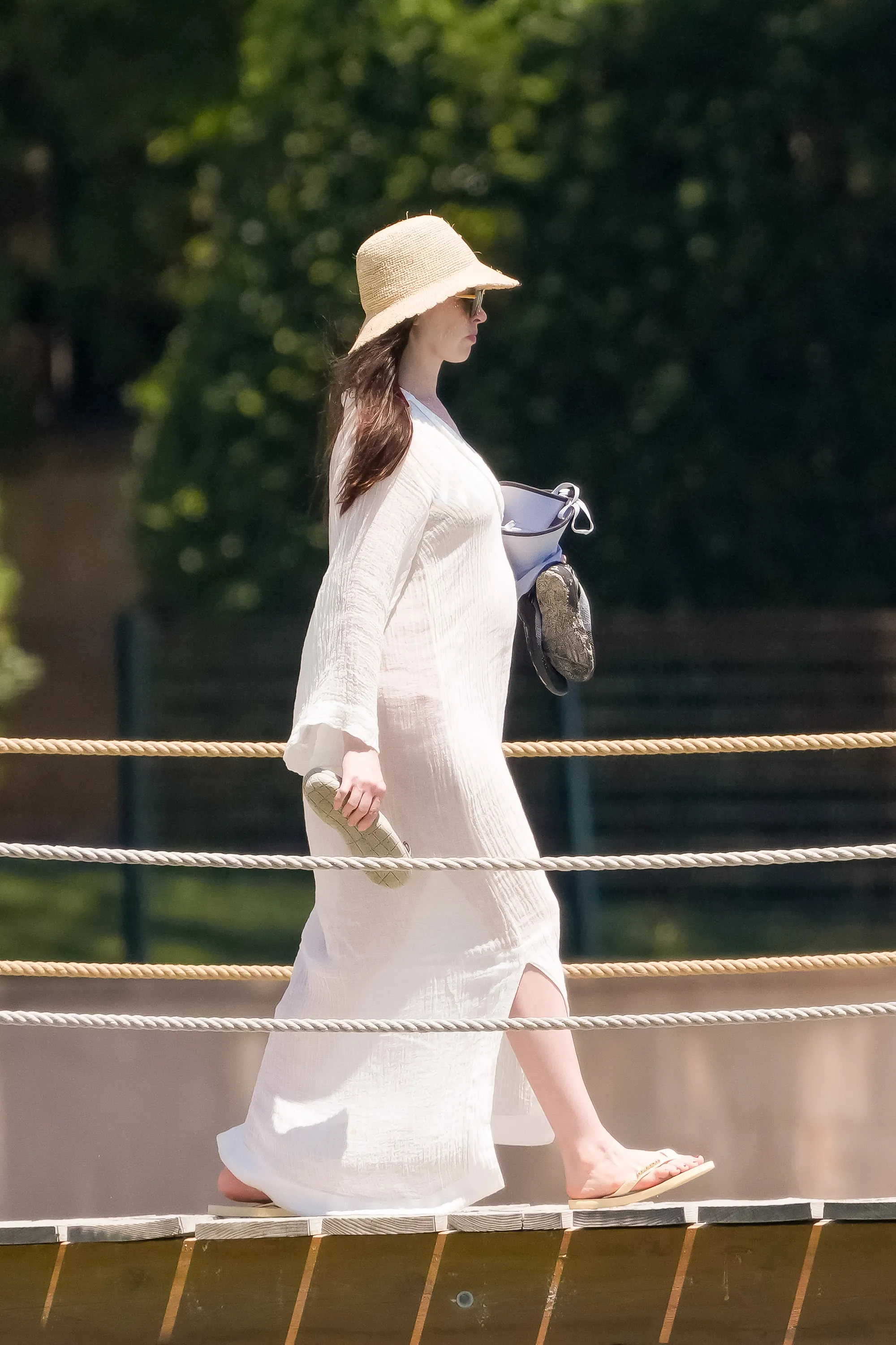 Anne Hathaway, wearing a white cover-up and straw hat, walks on a boardwalk, revealing a baby bump.