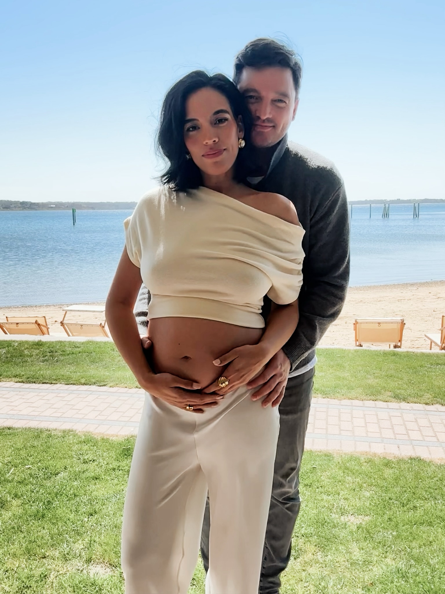 Daniell Olivera and her partner embracing, with her hands and his hands on her pregnant belly, standing near a beach.