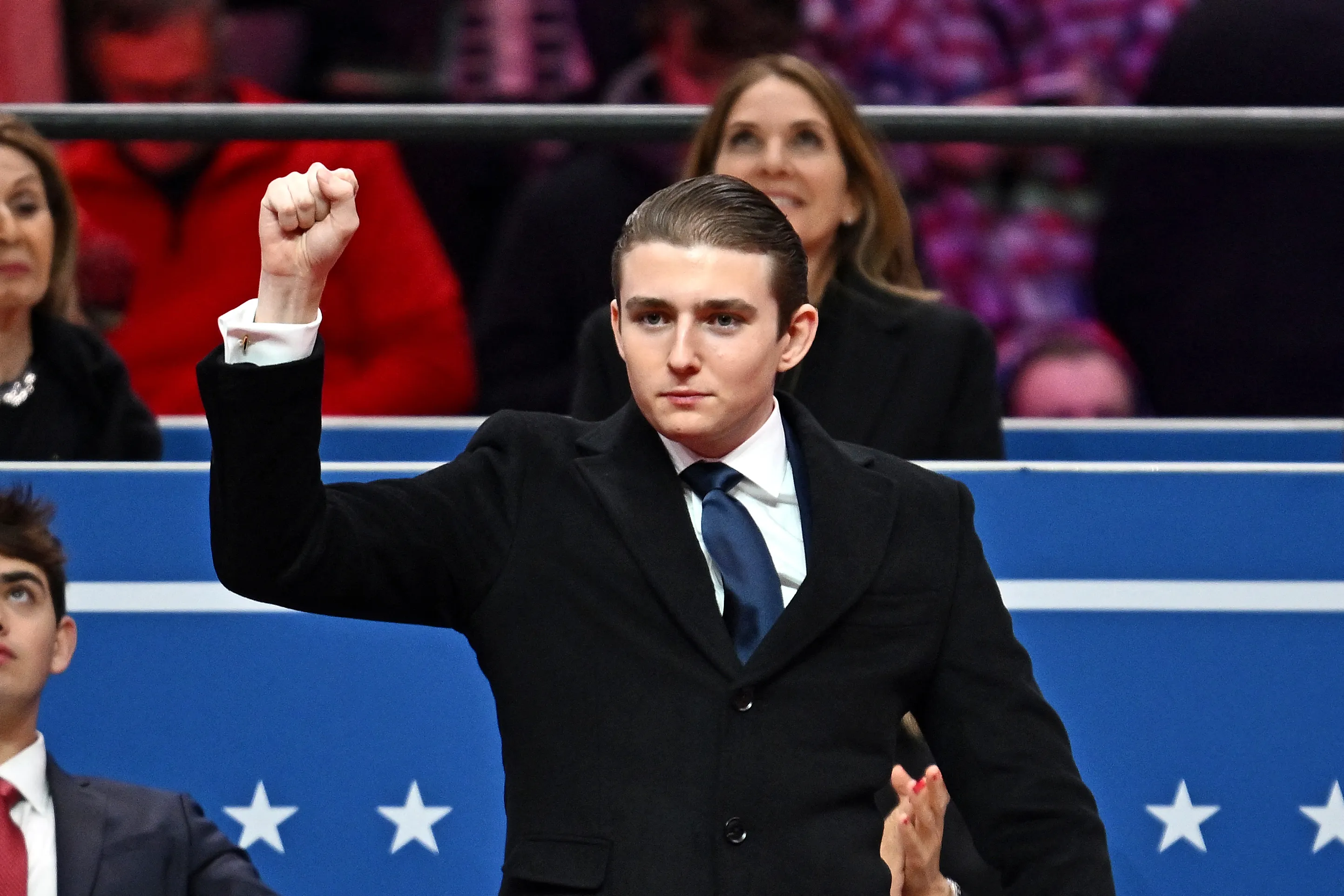 Barron Trump gestures with a raised fist while wearing a black coat, white shirt, and blue tie, with a woman looking on behind him.