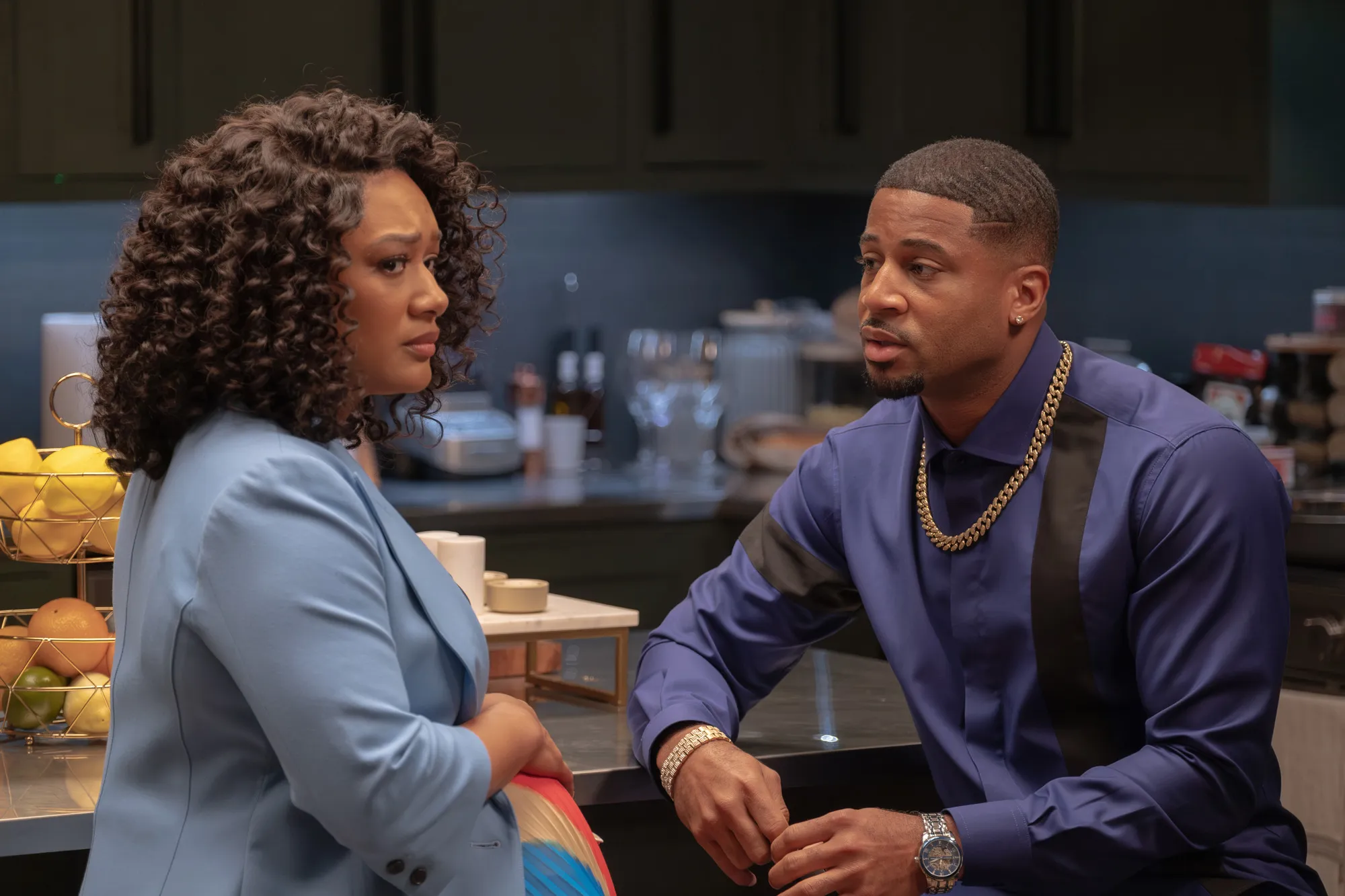 A woman with curly hair and a man in a blue shirt talking to each other in a kitchen.