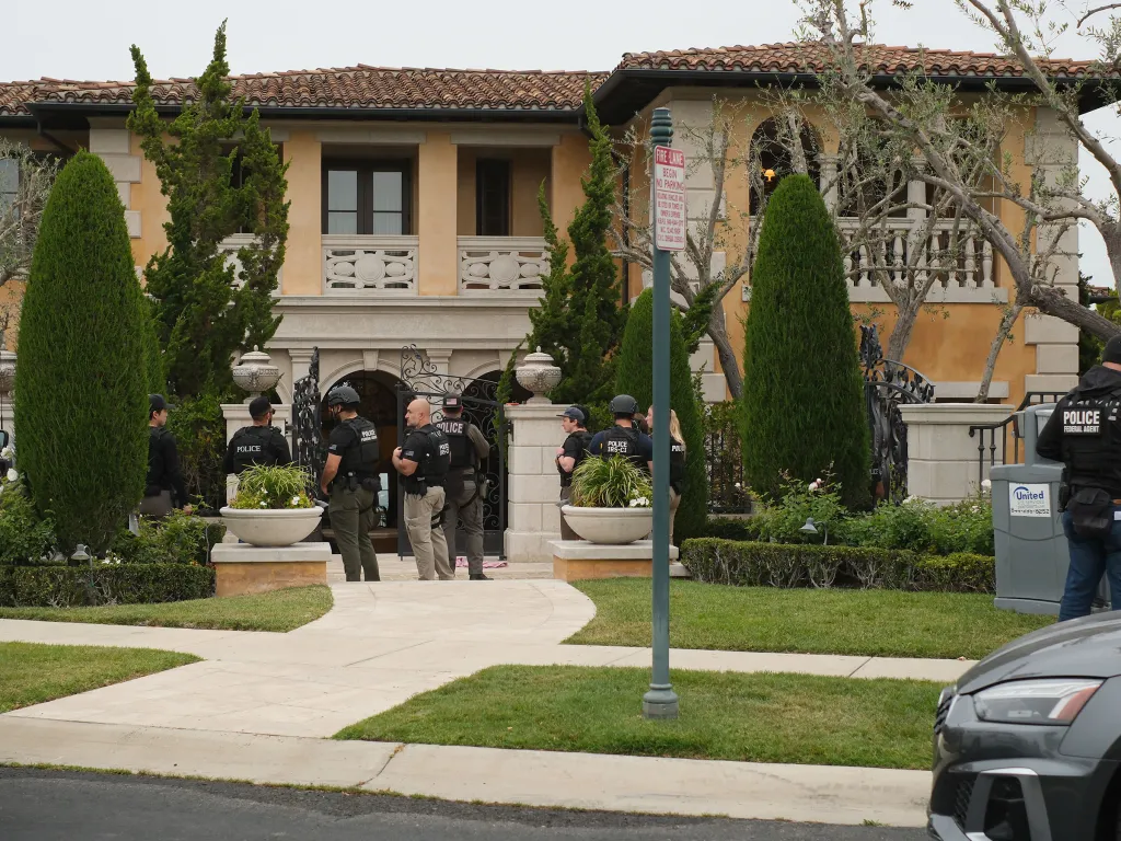 IRS agents and other police officers gather outside a large, upscale home with a red-tiled roof and light-colored stucco walls.