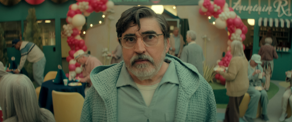 Alfred Molina as Sam in The Boroughs, wearing a light blue knitted jacket and glasses, looking directly at the camera with a surprised expression.