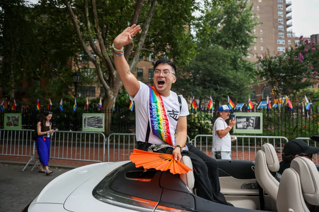 Bowan Yang, a Grand Marshal for the 2026 NYC Pride March, sits in a convertible, wearing a rainbow sash and holding an orange fan, as he waves to the crowd.