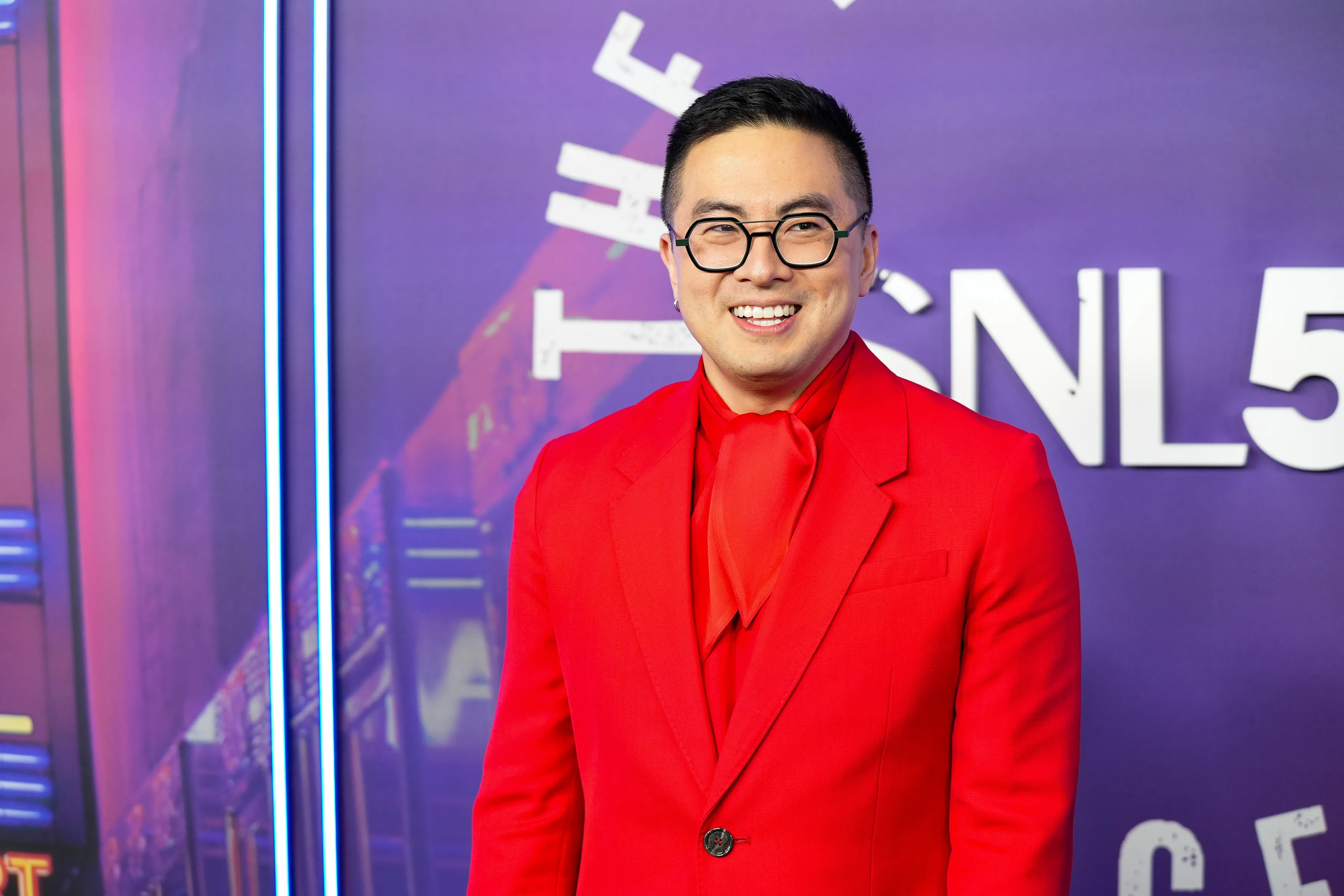 Bowen Yang smiling in a red suit and scarf against a purple background with white text.