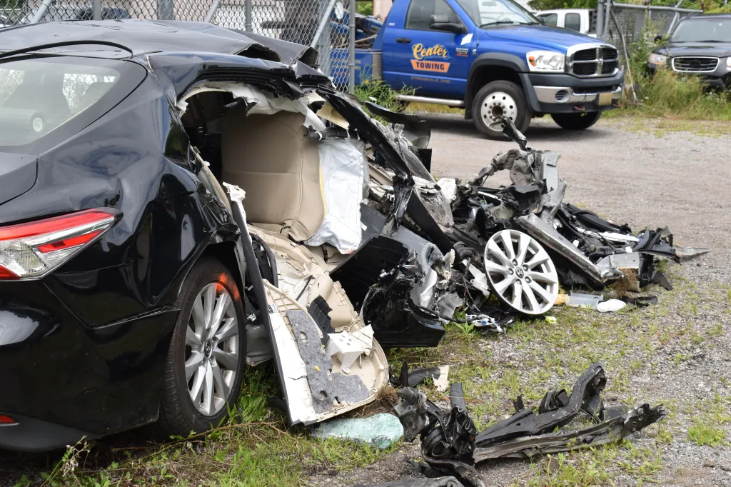 Remains of a severely crashed black car with debris scattered on the ground.