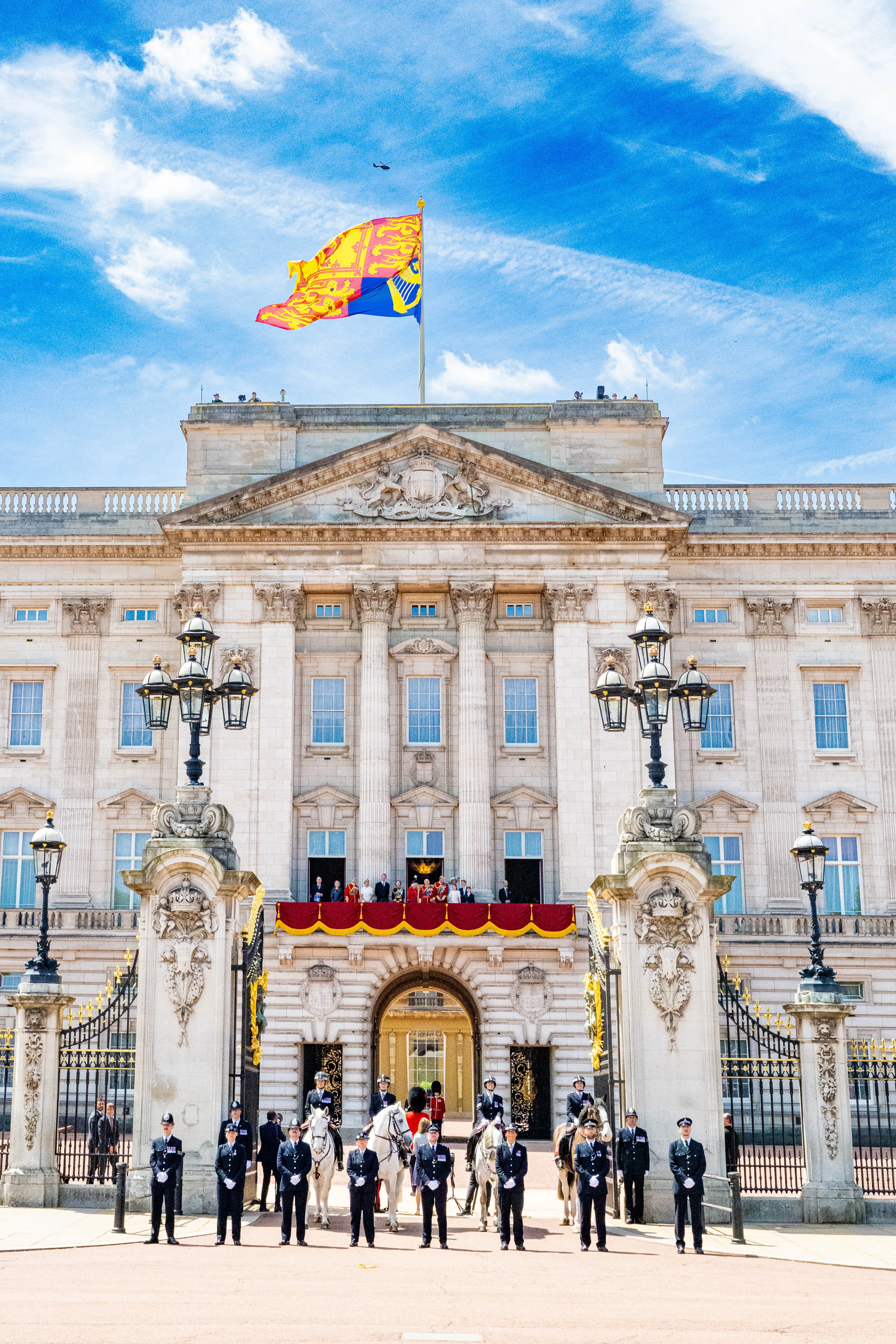 The Royal Family, including King Charles III and Queen Camilla, stand on the Buckingham Palace balcony during the Trooping the Colour ceremony, with guards on horseback and on foot below.